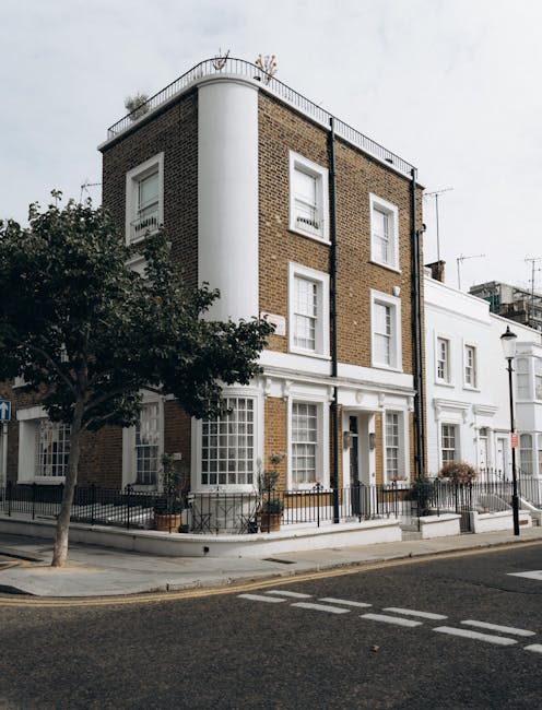 A multi-storey Victorian residential building situated on a street corner in Notting Hill, London, featuring a white corner column and a brown brick façade with white-framed sash windows. The ground level has a white-painted entrance with a small black metal fence and potted plants along the pavement. In front of the building, a black van with an open rear door is parked on the street, indicating an ongoing home relocation process by Man with Van Notting Hill. The street is lined with a tree, street lamps, and other white terrace houses. The image emphasizes careful handling during furniture transport and loading, consistent with professional removal services aimed at preventing damage to delicate Victorian floors and interior features, which are often part of house removals and packing and moving operations.