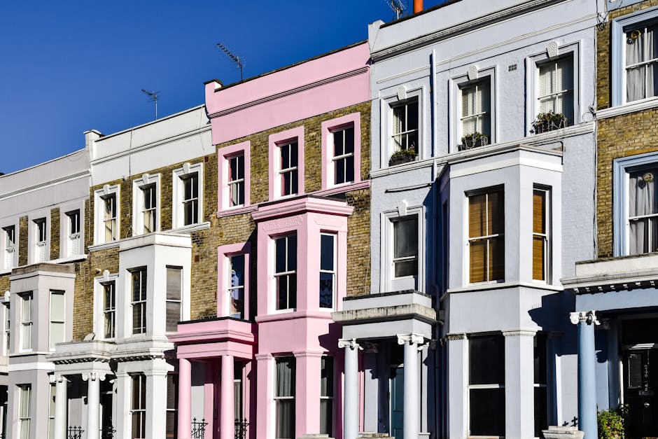 Photograph of a row of Victorian-style terraced houses in Notting Hill, London, showing detailed facades with brickwork and sash windows. The buildings feature a mix of colours, including white, pale pink, and light blue, with some bay windows and small flower boxes on the windowsills. A clear blue sky is visible above, and the image captures the exterior environment with natural sunlight. This setting depicts typical residential properties where house removals and relocation services by Man with Van Notting Hill are often required, as part of a furniture transport or home relocation process. The image emphasizes the importance of careful handling and planning in moving activities, especially in preserving the integrity of historic Victorian architecture during the loading and transport phases.