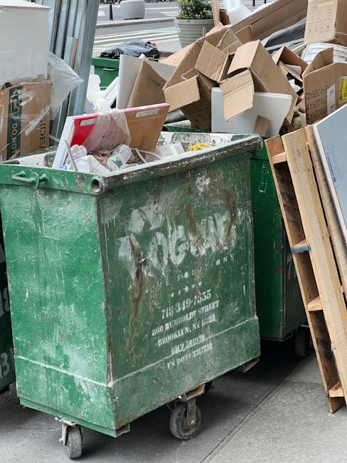 A large, green, industrial-sized waste skip filled with mixed household rubbish, including flattened cardboard boxes, plastic packaging, and small household items, situated on a street pavement in front of a residential property. The skip shows signs of wear with scratches and dirt, and is positioned near a building entrance with a wooden pallet placed beside it. Visible in the background are additional cardboard boxes and packaging materials, some stacked on top of each other, along with part of a vehicle passing by on the road. The image captures a typical scene during home relocation or furniture transport preparations, with the skip ready to be emptied or used for household waste clearance, illustrating the process of packing and sorting during house removals carried out by Man with Van Notting Hill.