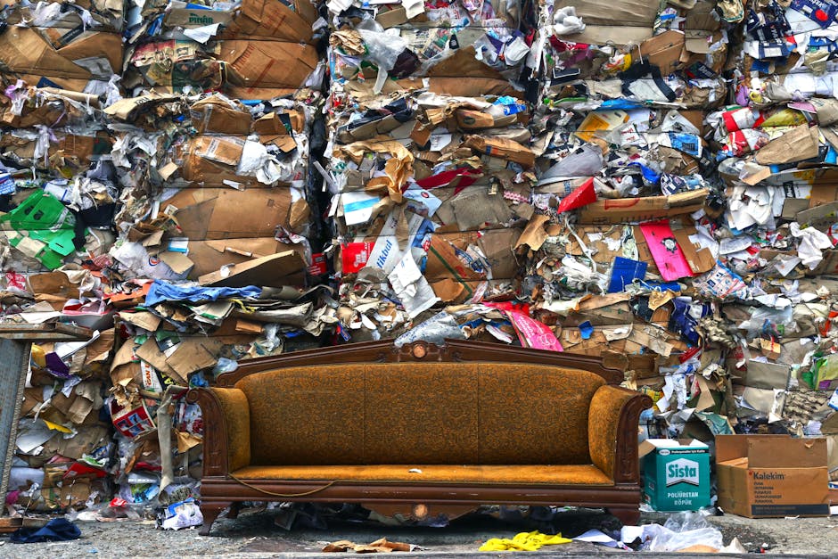 A brown vintage-style sofa with ornate wooden arms and a patterned fabric cushion is positioned on the pavement in front of a large pile of mixed waste materials. The waste includes crumpled cardboard boxes, torn paper, plastic packaging, and packaging tape, with some boxes stacked on top of each other and scattered across the area. The surrounding environment appears to be an outdoor space, possibly near a building or a waste collection site. Visible in the background are several open cardboard boxes, including one labeled 'Sista,' and a small yellow safety item lies on the ground in front of the sofa. The scene captures the process of waste removal or disposal, relevant to home relocation or clearing services by Man with Van Notting Hill, as part of packing and unloading during a move or waste disposal effort.