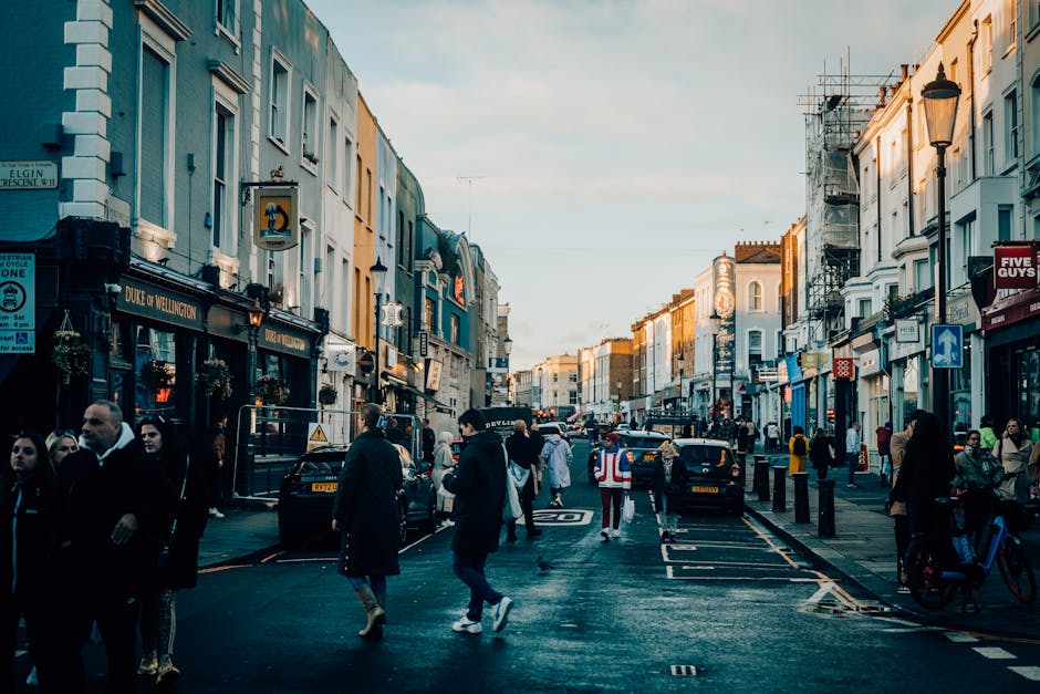 A busy street scene in Notting Hill W11 during daylight, with a mix of pedestrians walking along the pavement and crossing the road, some carrying bags or backpacks. Several parked cars are lining the side of the street, with a few vehicles in motion. On the left side, there are storefronts and cafes with signs, including one for Duke of Wellington, with a crowd gathered outside. The street is flanked by multi-storey buildings with light-colored facades, some under scaffolding or scaffolding structures indicating ongoing maintenance. Street lamps and road markings, including a bicycle lane, are visible. The scene depicts an active, urban environment suitable for home relocation or furniture transport planning, with conditions ideal for moving services such as loading and unloading of household items, furniture, or packing materials near the pavement and vehicles, as handled by companies like Man with Van Notting Hill.