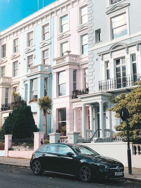 A black hatchback car is parked on a street in front of a row of tall, Victorian-style terraced houses painted in pastel shades of white, pink, and light blue, with decorative window surrounds and sma