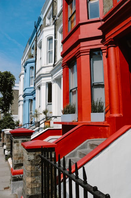 A black hatchback car is parked on a street in front of a row of tall, Victorian-style terraced houses painted in pastel shades of white, pink, and light blue, with decorative window surrounds and sma