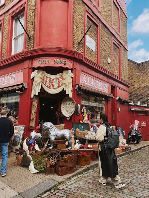 Exterior view of a red-brick building with ornate white decorations and signage indicating 'Alice's,' established in 1867, functioning as a shop or café in Notting Hill. Outside, there are various vintage and decorative items displayed on the pavement, including a large ceramic or stucco dog, ceramic ducks, and wooden crates with additional ornaments. Several people are visible; one person with a backpack is standing nearby, observing the display, while others are walking past. The atmosphere shows shop front windows with framed posters and signs, and the cobbled street with motor scooters parked against the building wall. The scene suggests a lively street scene during daytime, with natural lighting, and is relevant to the context of house or business relocations, potentially involving packing or moving activities in the area, as part of a professional removals service in Notting Hill.