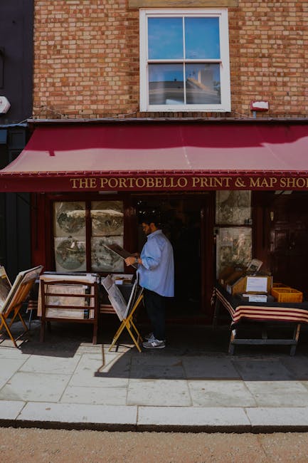 Exterior view of a red-brick building with ornate white decorations and signage indicating 'Alice's,' established in 1867, functioning as a shop or café in Notting Hill. Outside, there are various vintage and decorative items displayed on the pavement, including a large ceramic or stucco dog, ceramic ducks, and wooden crates with additional ornaments. Several people are visible; one person with a backpack is standing nearby, observing the display, while others are walking past. The atmosphere shows shop front windows with framed posters and signs, and the cobbled street with motor scooters parked against the building wall. The scene suggests a lively street scene during daytime, with natural lighting, and is relevant to the context of house or business relocations, potentially involving packing or moving activities in the area, as part of a professional removals service in Notting Hill.