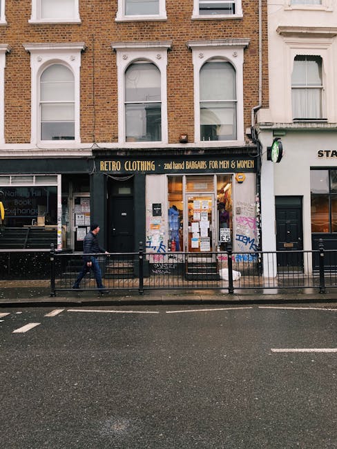 Image of a street view in Notting Hill featuring a retail shop named 'Retro Clothing' with a black sign and yellow lettering, offering second-hand bargains for men and women. The shop’s large front window displays various items and notices, with posters affixed to the window and door. The building has a brick façade with white-framed, arched windows above the shop. Outside, a person dressed in dark clothing is walking along the pavement, and a black metal railing separates the sidewalk from the road. The street appears wet, indicating recent rain, and there are no vehicles visible in this part of the street. The scene captures typical urban activity, relevant to house removals and moving logistics in Notting Hill, with the shop as a potential location for packing or retail furniture transport, aligned with home relocation and furniture moving services provided by Man with Van Notting Hill.