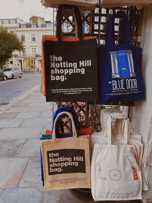 A display of eco-friendly reusable shopping bags hanging on a metal grid outside a property. There are four bags in total: a black bag with white text reading 'the Notting Hill shopping bag,' a blue bag with a white logo and text 'BLUE DOOR NOTTING HILL LODGE,' and two beige bags, one of which also features the same black text and logo. The bags are made of woven fabric and some have handles, with the black and beige bags positioned at the front and the blue bag hanging behind them. In the background, a street scene with parked cars, apartment buildings, and a sidewalk is visible under natural daylight. This image is relevant to house removals and moving services provided by Man with Van Notting Hill, illustrating packing and transportation materials used during a home relocation or furniture transport process.
