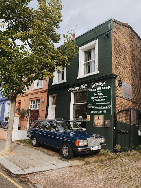 Image of a street view in Notting Hill featuring a retail shop named 'Retro Clothing' with a black sign and yellow lettering, offering second-hand bargains for men and women. The shop’s large front window displays various items and notices, with posters affixed to the window and door. The building has a brick façade with white-framed, arched windows above the shop. Outside, a person dressed in dark clothing is walking along the pavement, and a black metal railing separates the sidewalk from the road. The street appears wet, indicating recent rain, and there are no vehicles visible in this part of the street. The scene captures typical urban activity, relevant to house removals and moving logistics in Notting Hill, with the shop as a potential location for packing or retail furniture transport, aligned with home relocation and furniture moving services provided by Man with Van Notting Hill.