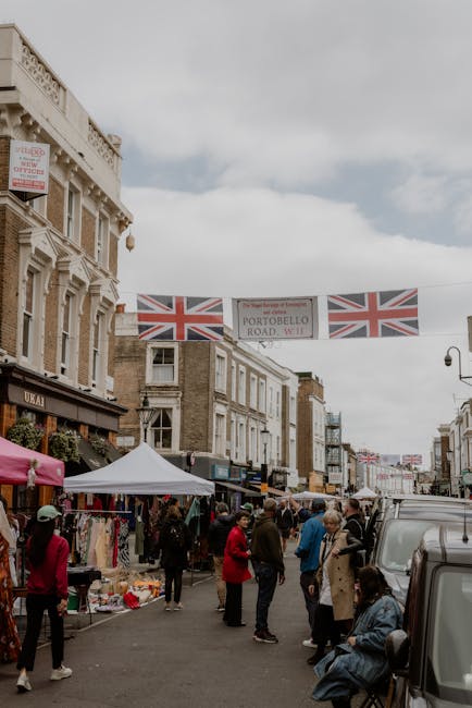 A bustling street scene in Notting Hill during a market day, with multiple people walking and browsing vendor stalls set up along the sidewalk. The stalls are covered with colorful canopies, displaying various clothing, accessories, and other goods. In the foreground, a person wearing a pink jacket and a hat is walking past a stall, while another person in a beige coat sits on a bench near the sidewalk. Behind the stalls, there are rows of traditional Victorian-style terraced buildings with brick facades, some displaying British Union Jack flags and promotional signs. A grey van is parked on the right side of the street, and overhead, decorative flags featuring the Union Jack are strung across the street, adding a festive atmosphere. The scene captures the lively character of a typical day in Notting Hill, which often involves home relocation, furniture transport, and packing and moving activities managed by companies like Man with Van Notting Hill, who may be involved in local removals services.
