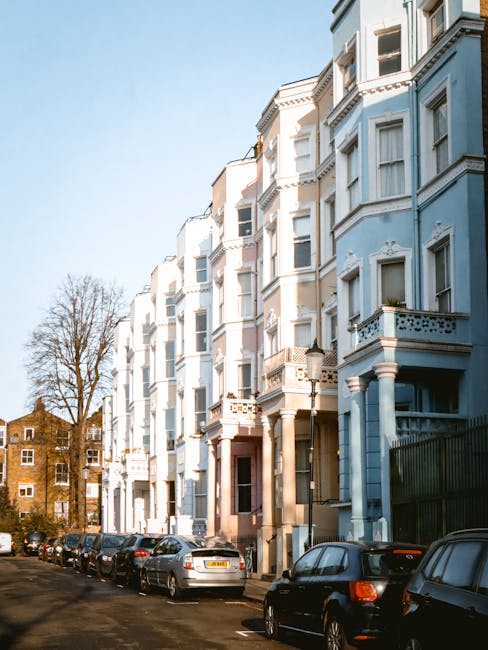 A row of white Victorian-style terraced houses with ornate detailing and bay windows, situated along a residential street with parked cars lining the pavement. The buildings are multi-storey, featuring decorative balconies and stone columns at the entrance. In the foreground, a moving company, Man with Van Notting Hill, is engaged in a home relocation process. A large wooden wardrobe wrapped in protective blankets and secured with plastic wrap is being carried by two movers, with one lifting it onto a trolley or dolly. Nearby, several cardboard boxes and packed items are visible, suggesting packing and moving preparations. The scene occurs during daylight with clear blue skies, and the surrounding environment includes leafless trees and additional residential buildings in the background. This image illustrates the logistics and efficient furniture transport involved in house removals within the W11 area, supporting the services provided by Man with Van Notting Hill in their moving and packing operations.