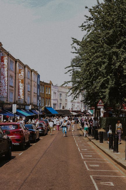 A bustling street scene in Notting Hill during a market day, with multiple people walking and browsing vendor stalls set up along the sidewalk. The stalls are covered with colorful canopies, displaying various clothing, accessories, and other goods. In the foreground, a person wearing a pink jacket and a hat is walking past a stall, while another person in a beige coat sits on a bench near the sidewalk. Behind the stalls, there are rows of traditional Victorian-style terraced buildings with brick facades, some displaying British Union Jack flags and promotional signs. A grey van is parked on the right side of the street, and overhead, decorative flags featuring the Union Jack are strung across the street, adding a festive atmosphere. The scene captures the lively character of a typical day in Notting Hill, which often involves home relocation, furniture transport, and packing and moving activities managed by companies like Man with Van Notting Hill, who may be involved in local removals services.