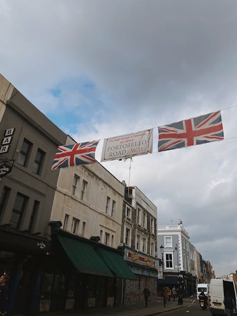 A street scene in Notting Hill showing a banner overhead with Union Jack flags on either side and a central sign reading 'Portobello Road W11'. The banner is strung across the road between buildings, suggesting a local event or market. Below, there are several shops with awnings, including one with a green canopy, and pedestrians walking along the pavement. A white van is parked nearby, indicating the presence of transportation for a house removal or delivery service, as associated with professional removals in Westbourne Grove. The sky above is partly cloudy, creating a typical urban atmosphere supportive of moving and packing activities in the area.