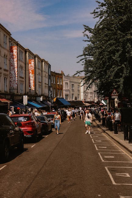 A street scene in Notting Hill showing a banner overhead with Union Jack flags on either side and a central sign reading 'Portobello Road W11'. The banner is strung across the road between buildings, suggesting a local event or market. Below, there are several shops with awnings, including one with a green canopy, and pedestrians walking along the pavement. A white van is parked nearby, indicating the presence of transportation for a house removal or delivery service, as associated with professional removals in Westbourne Grove. The sky above is partly cloudy, creating a typical urban atmosphere supportive of moving and packing activities in the area.