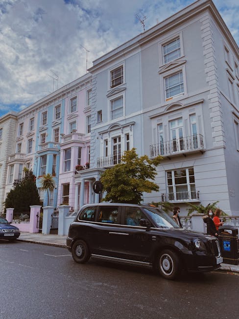 A black London-style taxi parked on a residential street in front of a white, multi-story Victorian townhouse during daytime. The townhouse has decorative cornices, large sash windows, and small balconies with black wrought iron railings. The building's façade displays detailed architectural moldings and is surrounded by greenery, including a prominent tree near the sidewalk. The street has a mix of parked cars and pedestrians, some of whom are near a trash bin, and the sky is partly cloudy. This scene illustrates a typical urban setting suitable for house removals and furniture transport, with the street likely used for loading and unloading during a home relocation, consistent with services provided by Man with Van Notting Hill.