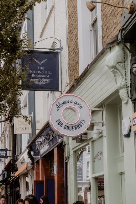 A two-storey brick building with a dark green facade housing Nothing Hill Garage, with a white sign displaying the business name and contact details, situated on a residential street. In front of the building, a vintage blue station wagon with a roof rack is parked on the pavement, partly covering a small gate and entrance. There is a large tree with green leaves in front of the building, casting shadows on the pavement and partially obscuring the upper windows. The building exterior includes white-framed windows on both floors, with the ground-floor window reflecting surrounding houses. Visible items related to house removals and home relocation include cardboard boxes and packing materials stacked near the entrance or inside, but they are not directly seen in this image. The scene captures a calm, suburban environment during daylight, with a focus on the building’s entrance and the parked vehicle, illustrating a typical setting for local moving and furniture transport services in Notting Hill, as part of [COMPANY_NAME]'s residential relocation support.